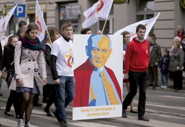 jóvenes participantes de la JMJ de Cracovia caminan por la calle con banderas y un cartel con la imagen de san Juan Pablo II